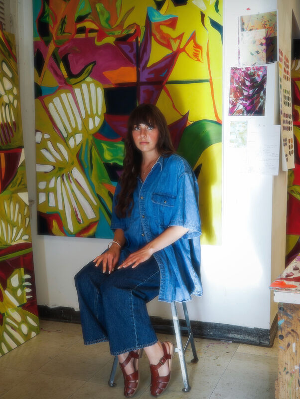 Mia Baker seated in her studio, surrounded by three large-scale Terrarium oil paintings in vivid reds, yellows, cyans, and greens.